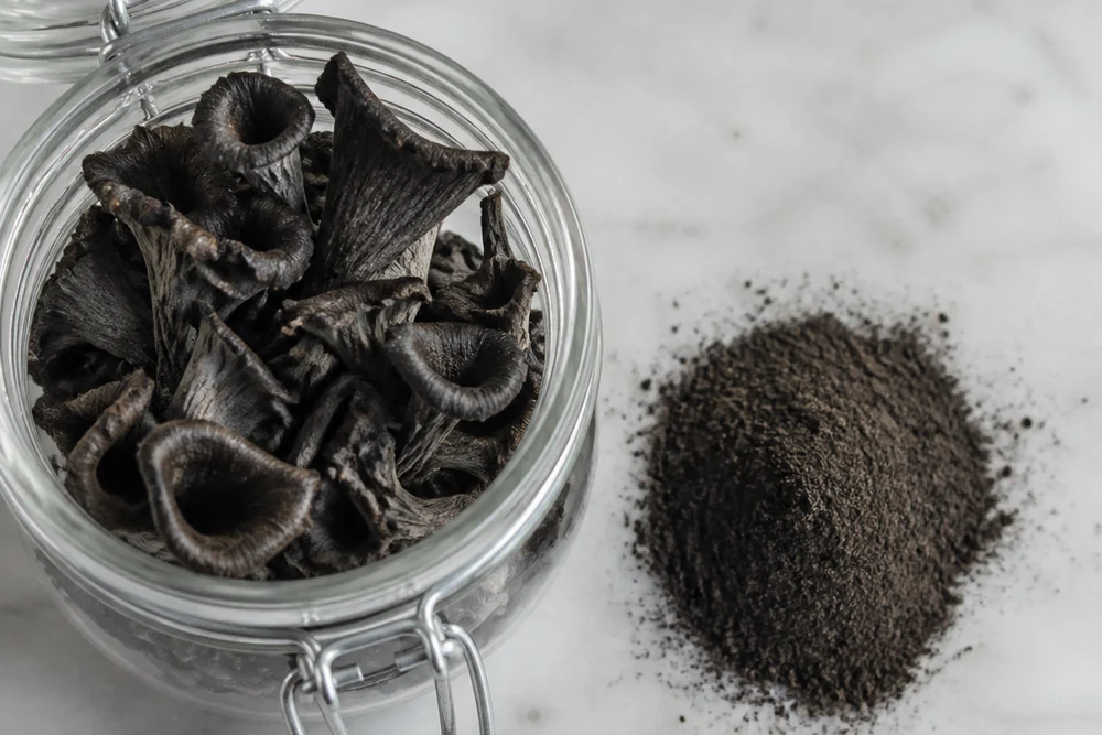 Dried black trumpet mushrooms in a glass jar beside a small heap of the same mushrooms ground to a dark powder