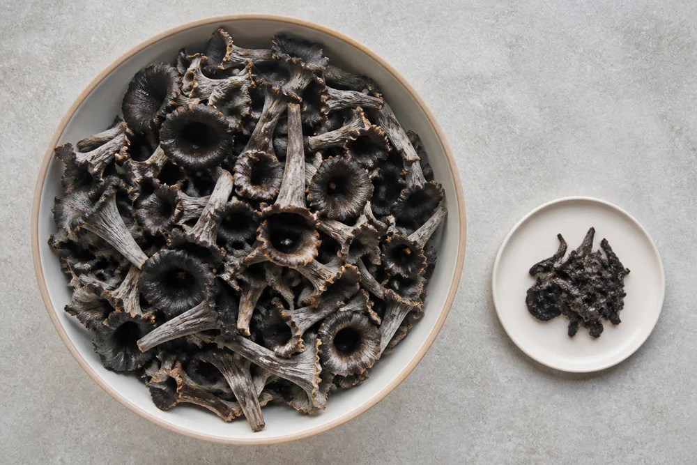 A large bowl of fresh black trumpet mushrooms beside a very small pile of the same mushrooms after cooking