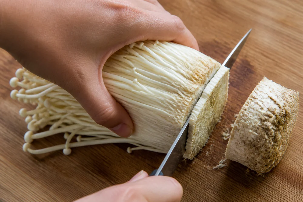 Hands cutting the dense compressed base off a bundle of enoki mushrooms on a wooden board with the discarded root plug beside the separated strands