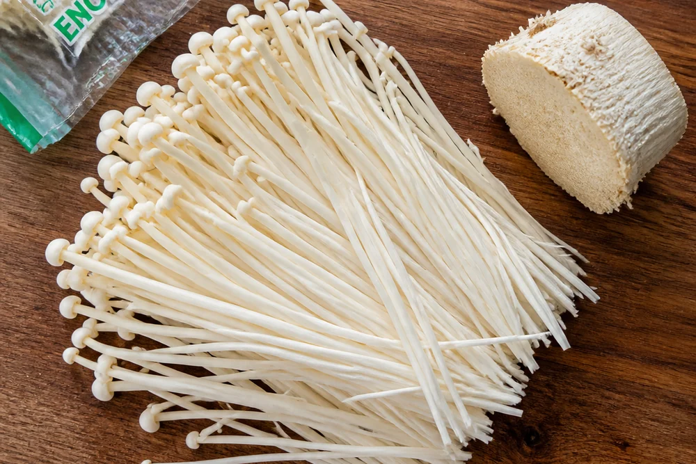 A pack of white enoki mushrooms opened on a board with the dense base plug cut away and the strands fanned out loose