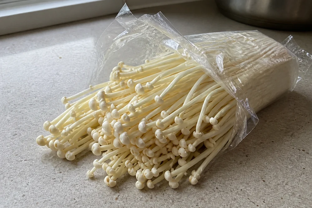 A vacuum-sealed plastic pack of white enoki mushrooms opened on a kitchen counter with the long thin stems and tiny caps clearly visible