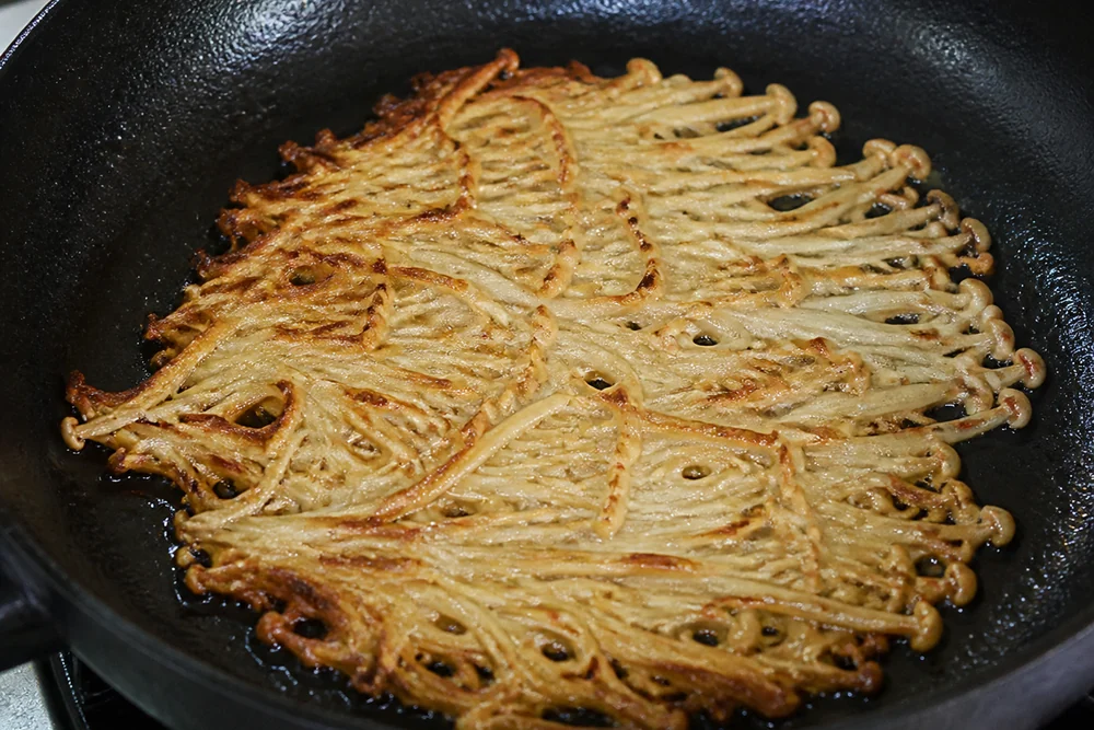 A tangle of enoki mushrooms browning in a hot skillet with the strands crisped and golden at the edges and pressed flat into a mat