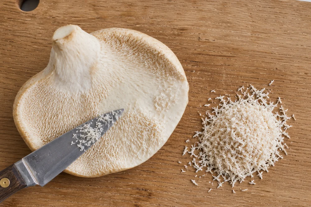 A hedgehog mushroom on a board with a knife scraping the spines off the underside, a small heap of loose spines beside it