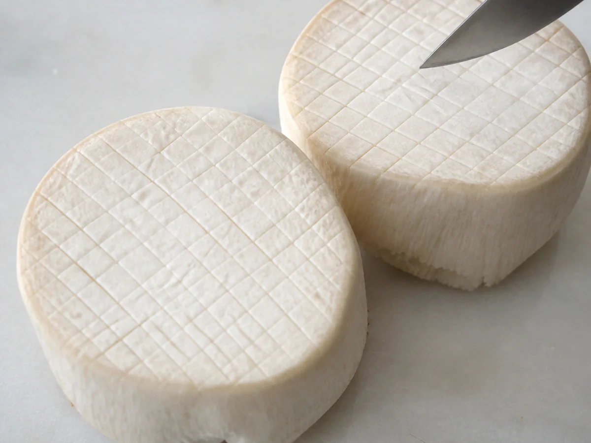 Two thick raw king oyster discs on a pale board, each scored across the cut face in a fine crosshatch pattern