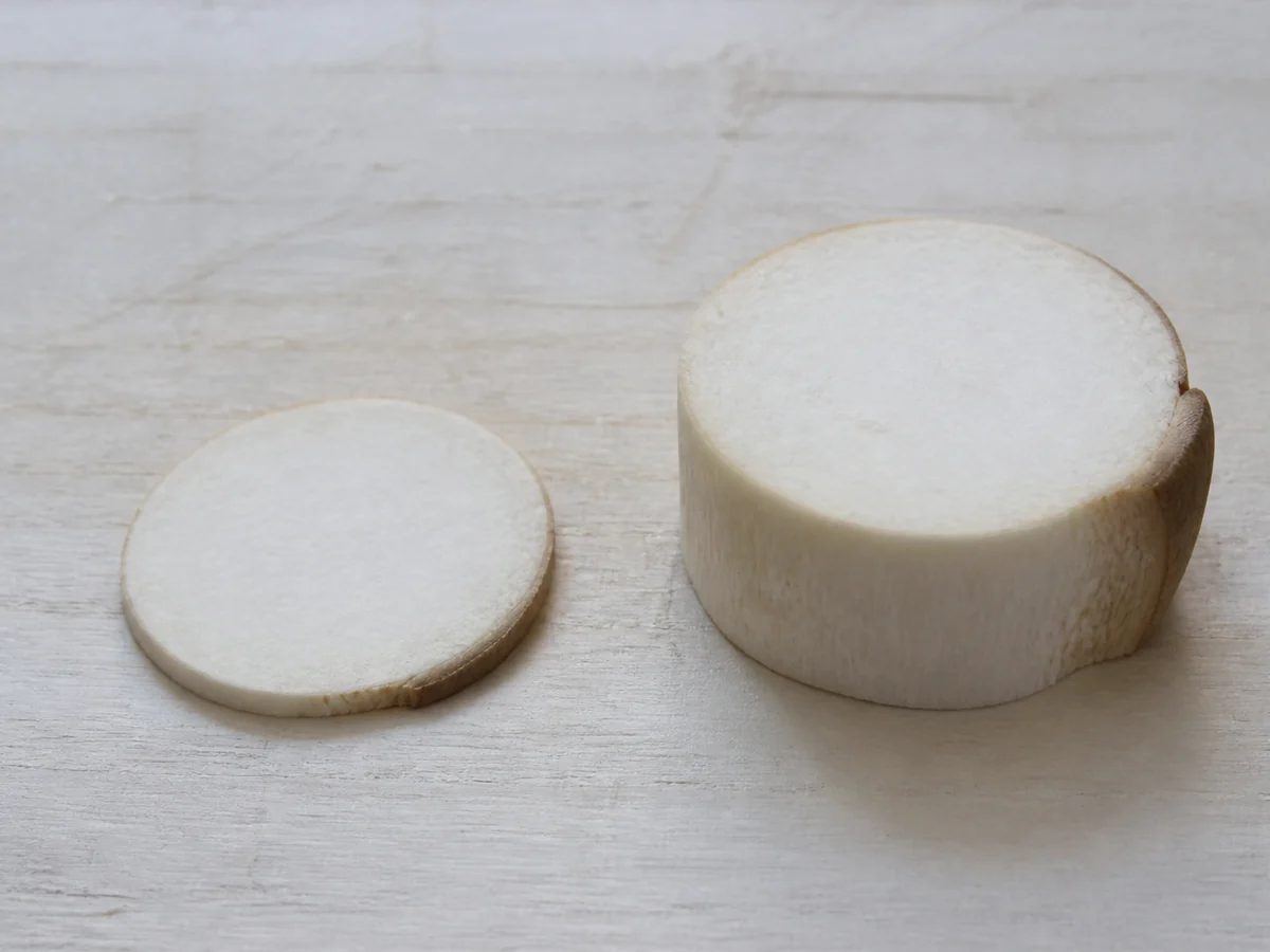 Two raw king oyster cut pieces side by side on a pale board, a very thin coin-thick slice beside a disc three times as tall
