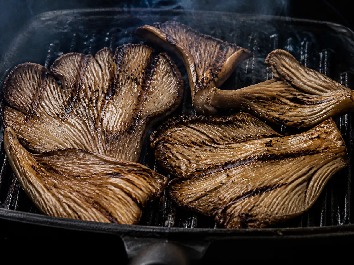 Large oyster mushroom pieces on a hot ridged grill pan with dark char lines across the pale fan-shaped caps