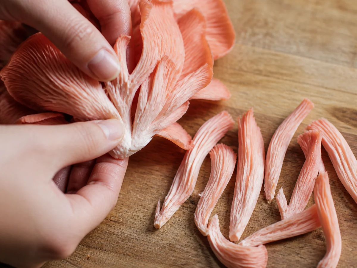 Pink oyster mushroom cluster being pulled apart by hand into thin strips on a wooden board with several strips already separated