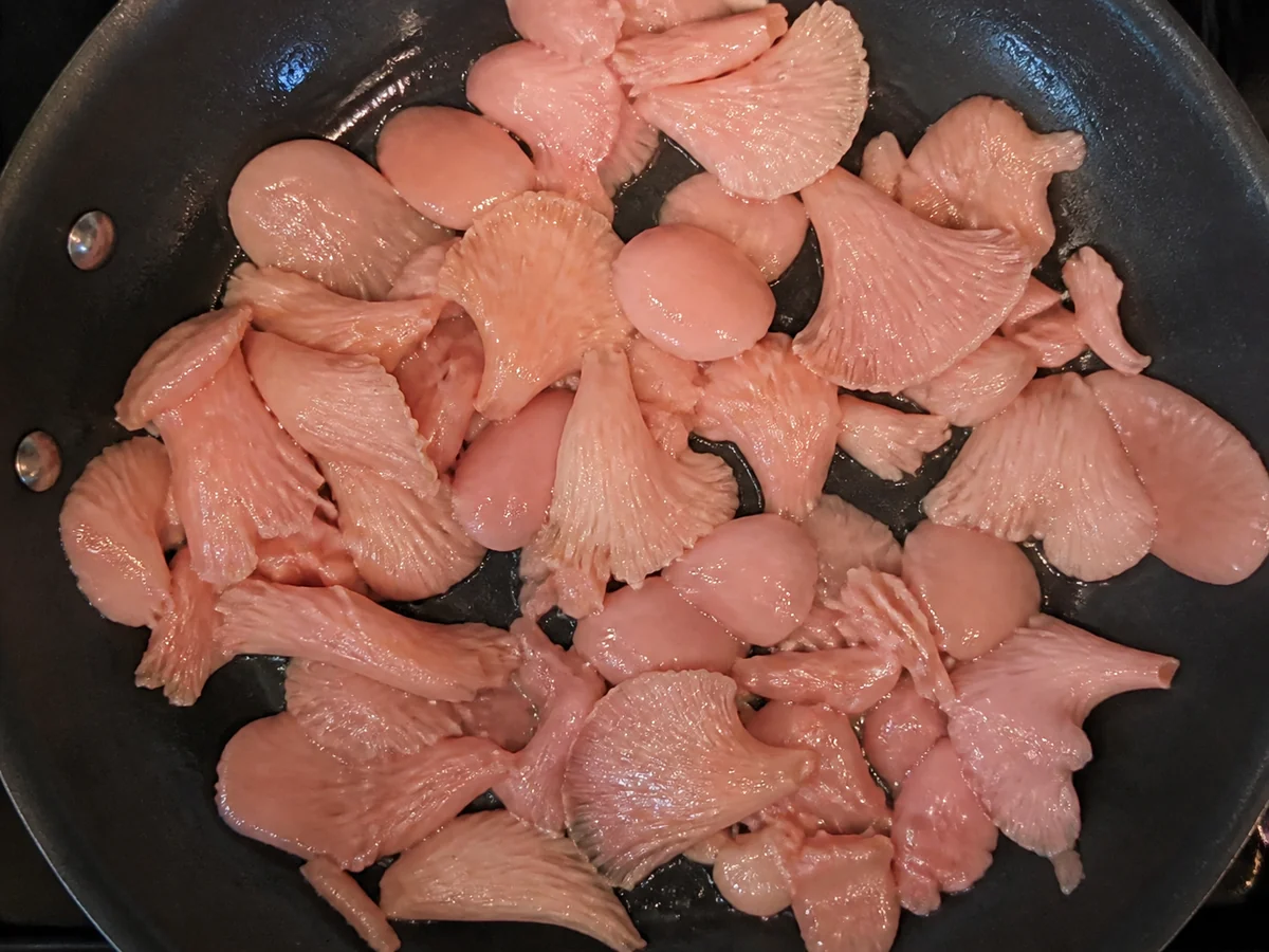 Pale pink oyster mushroom pieces in a pan barely cooked, still pink and floppy and shiny with moisture and no browning