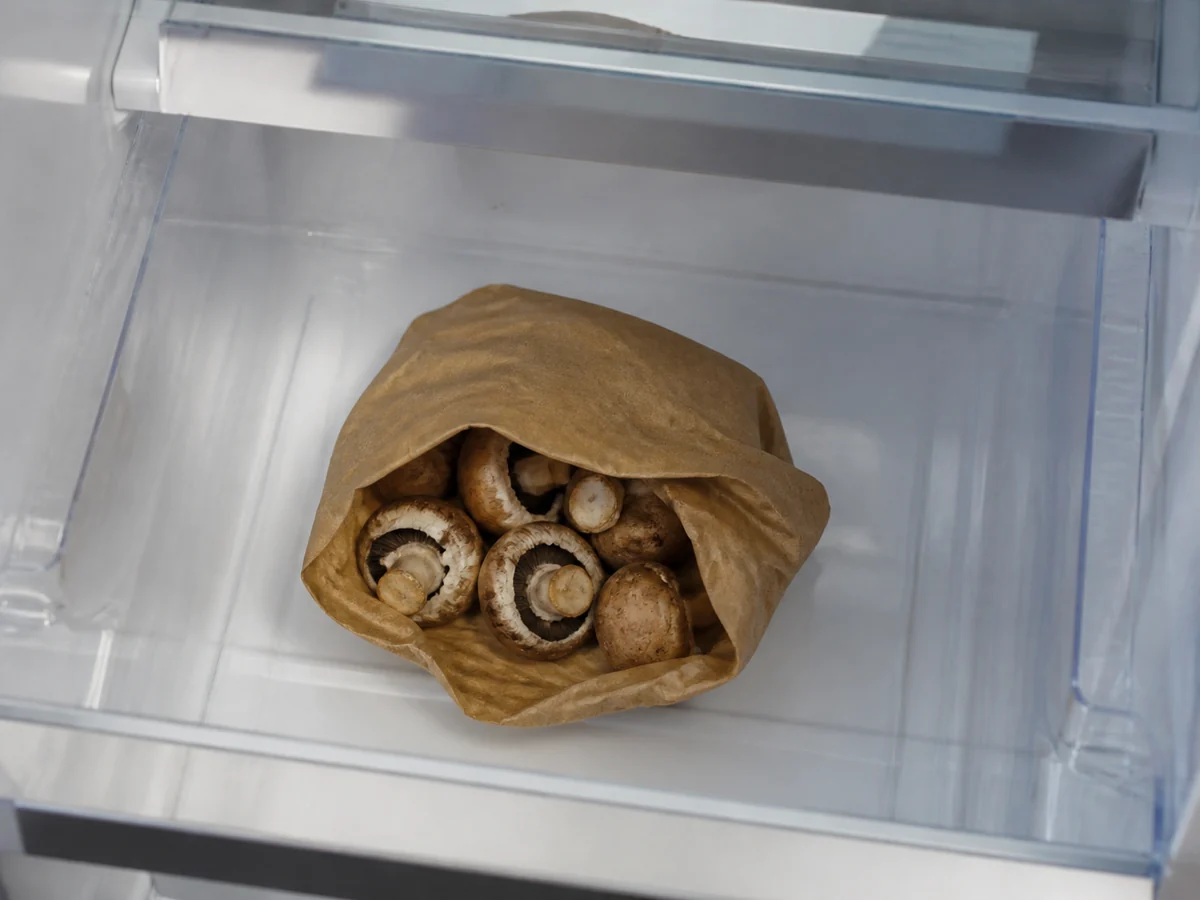 An open brown paper bag of cremini mushrooms lying in an otherwise empty refrigerator crisper drawer, the top of the bag open