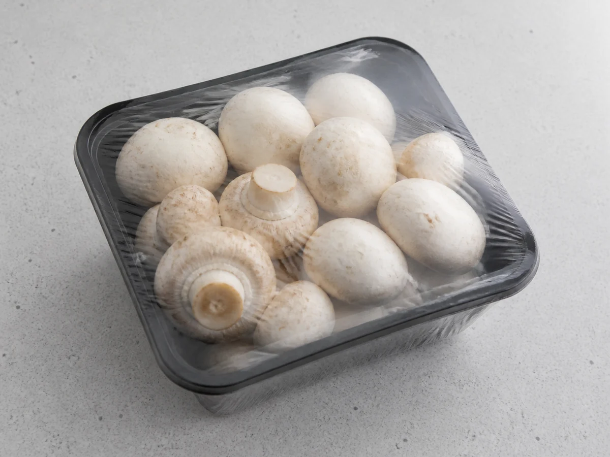 A supermarket punnet of fresh white button mushrooms with the clear plastic film still sealed over the top