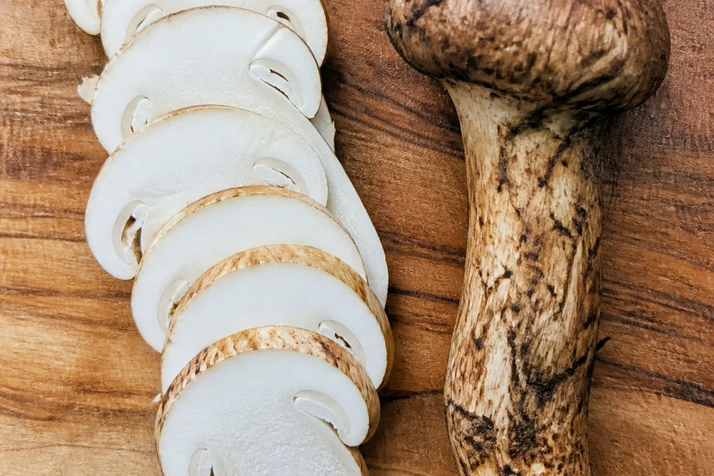 A matsutake mushroom sliced into thin pale rounds on a wooden board beside the intact stem