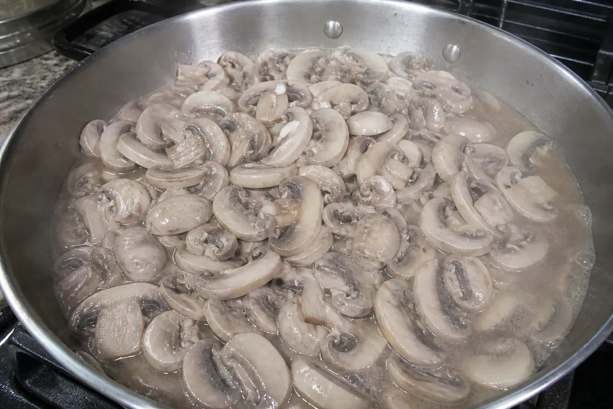 Pale sliced mushrooms crowded in a skillet with visible released water