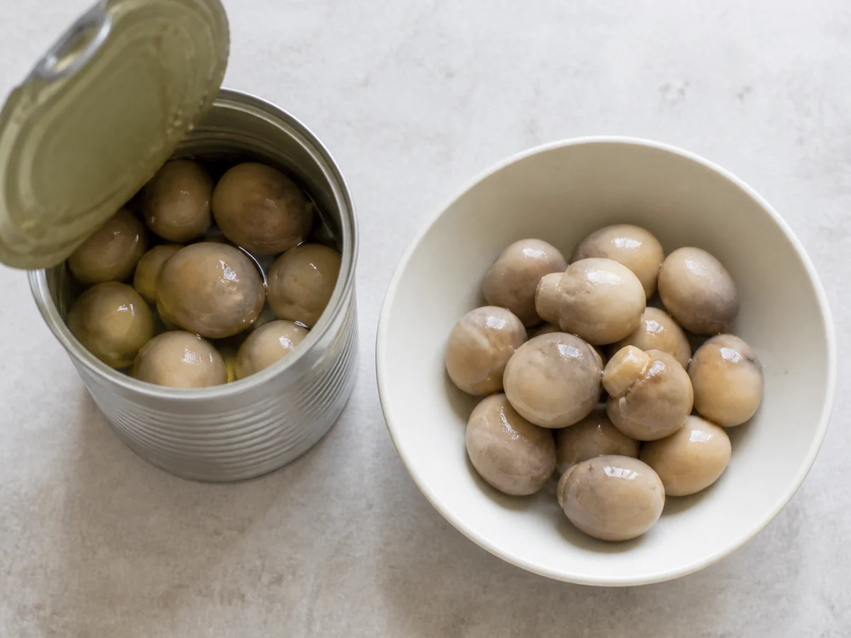 An opened can of straw mushrooms beside the drained pale ovals tipped into a white bowl