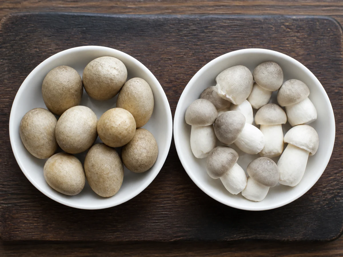 Two bowls side by side, one of smooth whole unpeeled straw mushrooms and one of peeled ones showing the cap and stem shape