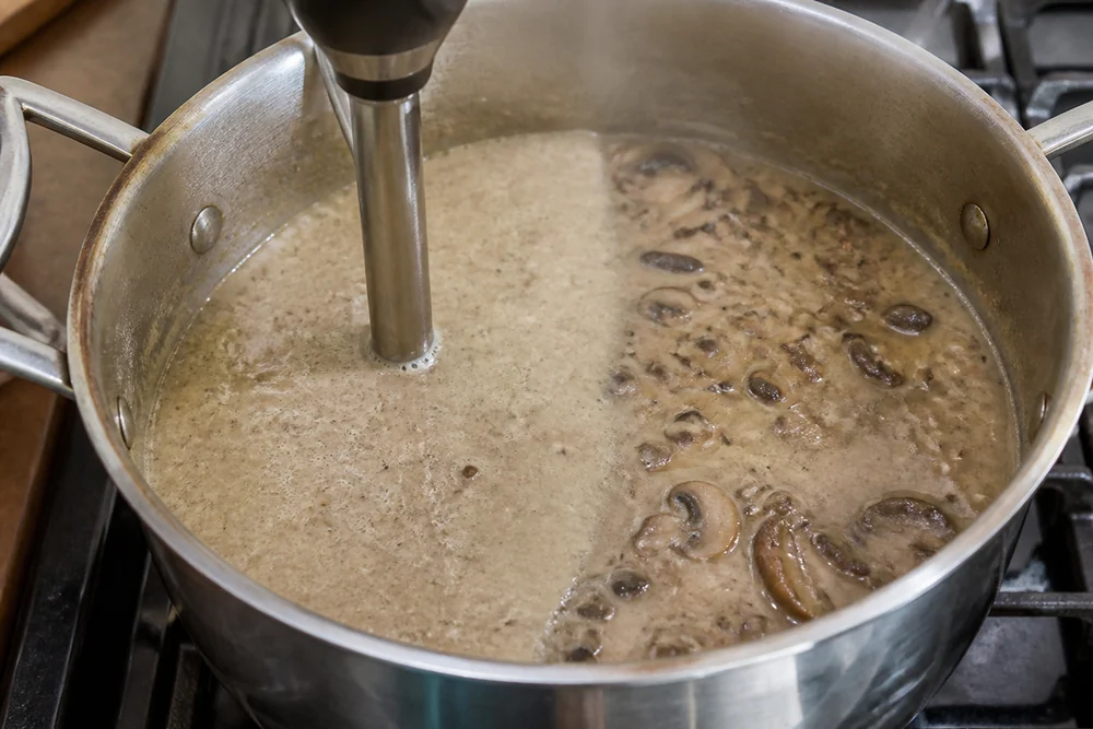 An immersion blender part-blending a pot of mushroom soup on a stove, half the pot smooth and half still showing mushroom pieces, steam rising