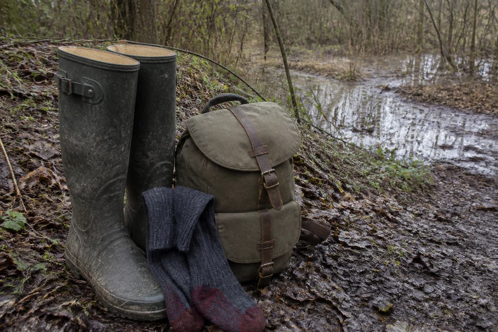 A pair of muddy waterproof boots and long socks beside a small backpack at the edge of wet woodland with damp leaf litter and standing water behind