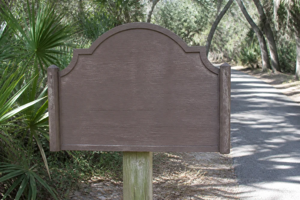 A brown Florida state park entrance sign beside a sandy road with cabbage palms and pines behind it