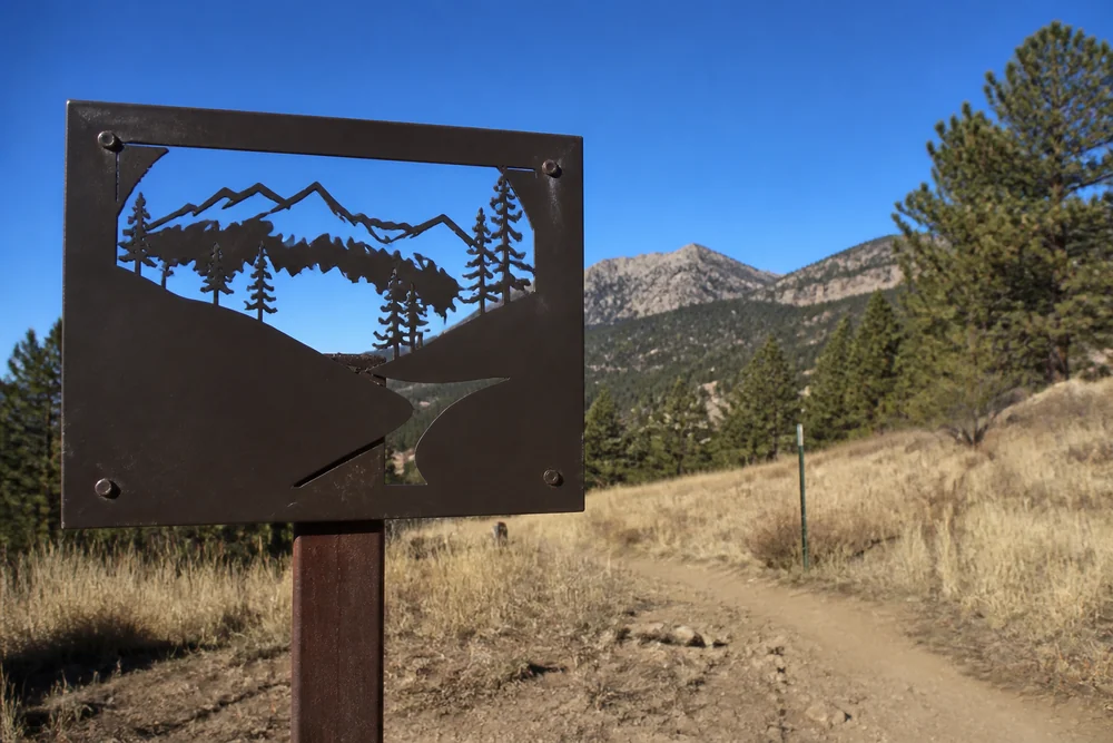 A metal trailhead sign on a post at the edge of a Colorado county open space area with dry grassland, scattered ponderosa pines and foothills behind