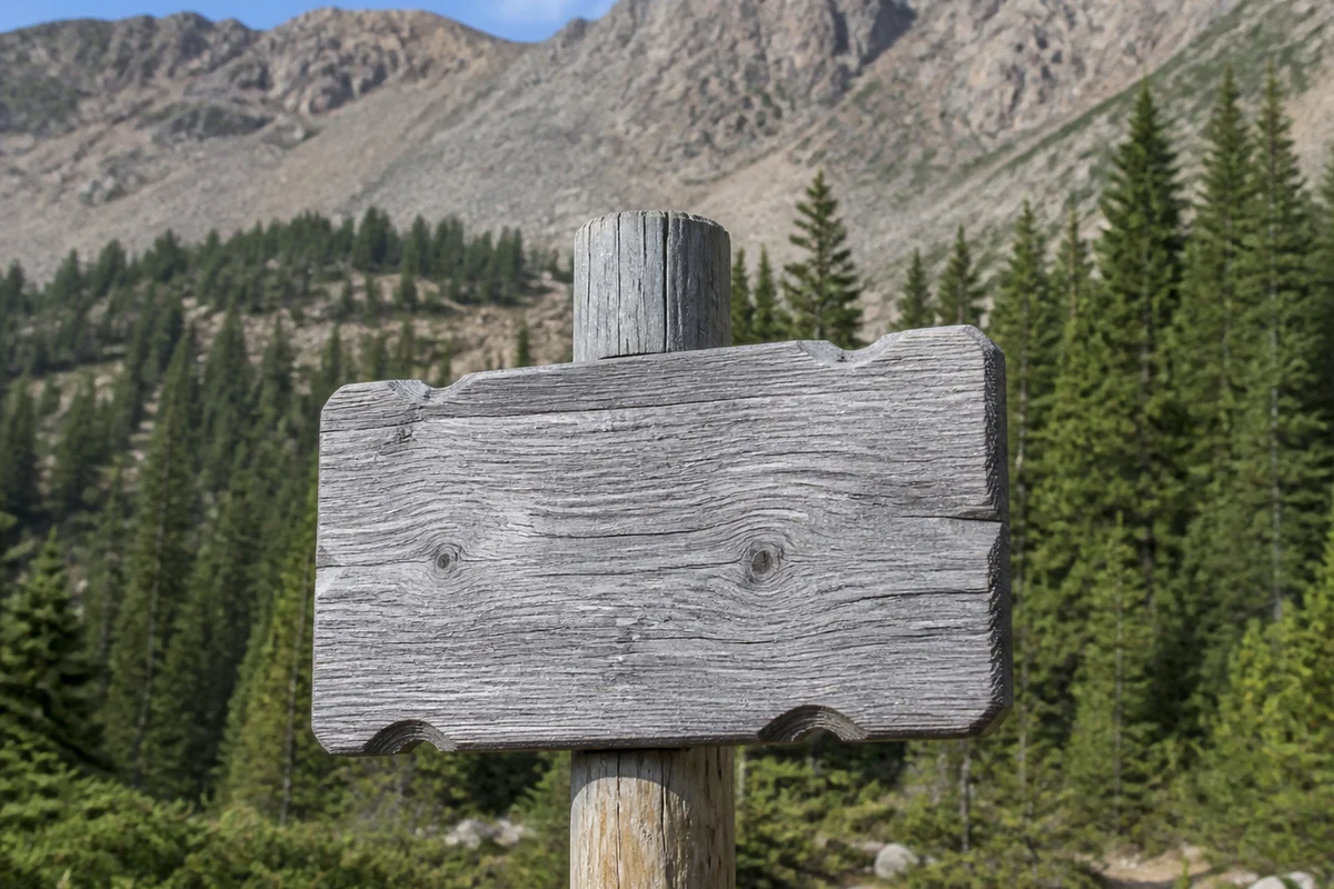 A routed wooden sign on a post at a Colorado high country trailhead with weathered bare timber, dense spruce behind and a rocky ridge above