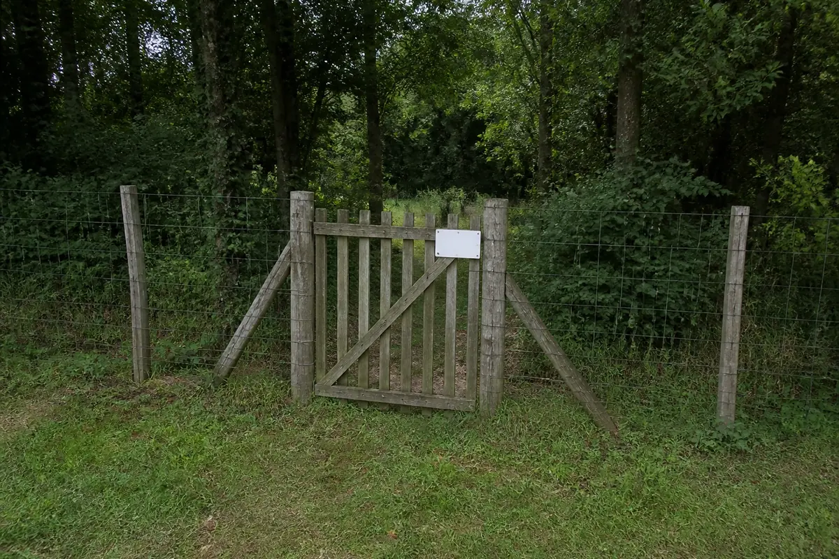 A wire fence and closed wooden field gate at the edge of private French woodland with a small blank enamel sign wired to the gate