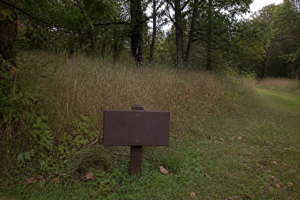 A small brown wooden sign on a low post at the edge of a quiet Wisconsin woodland clearing with tall grasses and oaks behind
