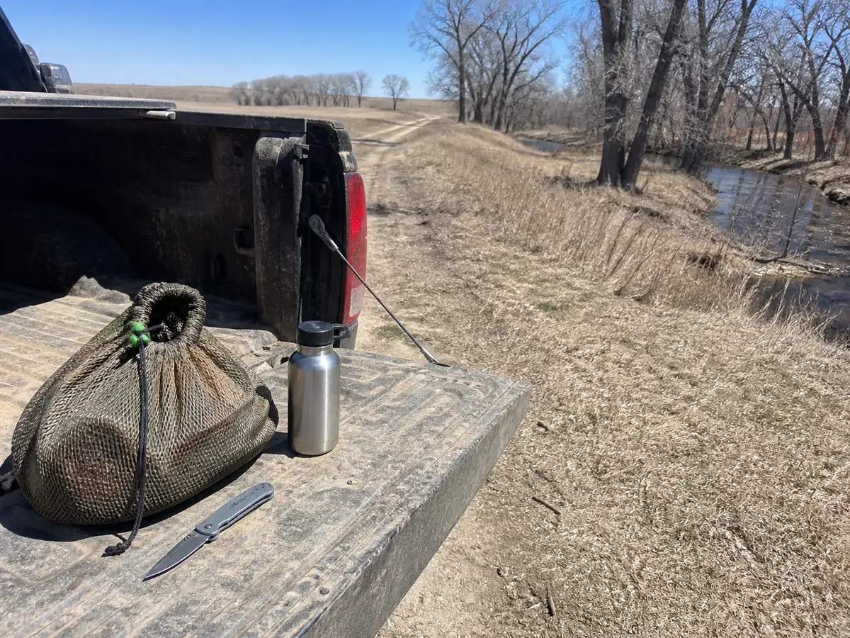 An open tailgate of a dusty pickup on a two track beside Kansas creek timber, with a mesh drawstring bag, a folding knife and a metal water bottle on it, open prairie and bare cottonwoods behind