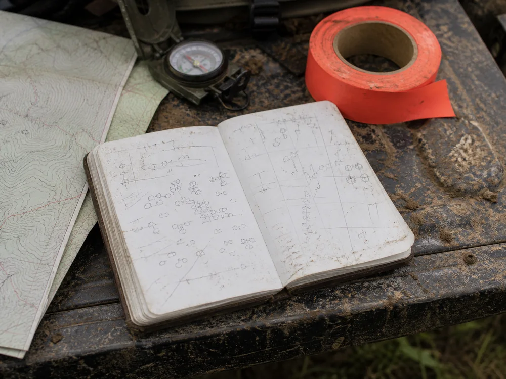 A weathered field notebook open on a truck tailgate beside a folded map, a compass and a roll of orange flagging tape