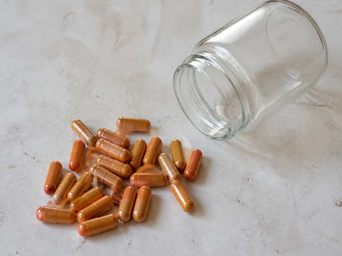 Small heap of plain orange brown capsules on a pale stone surface beside a plain glass jar lying on its side
