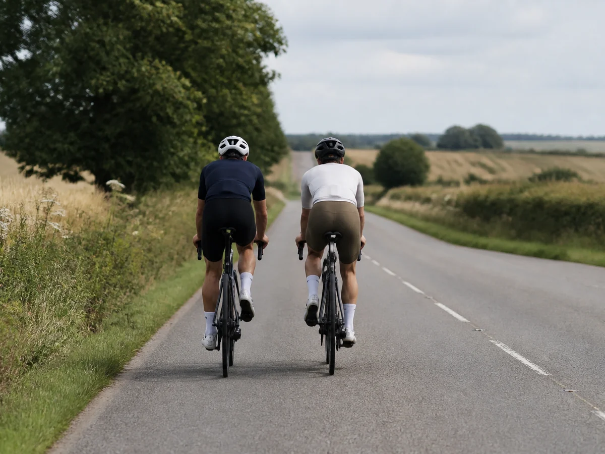 Two road cyclists riding side by side on an open country road in flat daylight, seen from behind at a distance