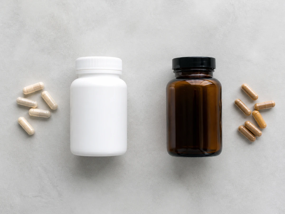 Two plain unlabeled containers side by side on a pale surface, a white pharmacy style bottle and an amber glass supplement jar, with loose capsules beside each