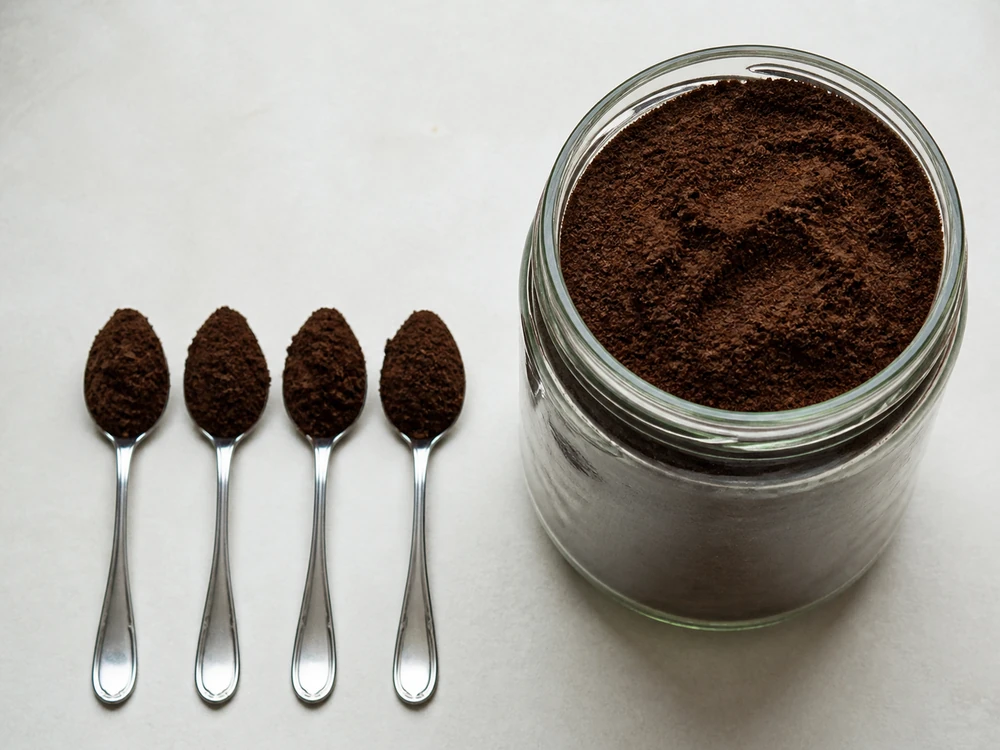 Four teaspoons of dark powder measured out on a pale surface beside a large jar holding the same daily amount multiplied over a month