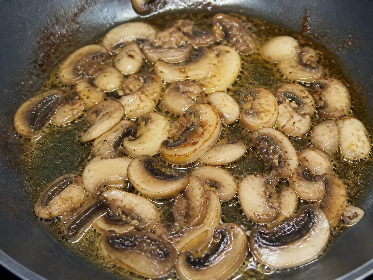 Sliced mushrooms saturated with butter and oil sitting in a shallow pool of fat in a frying pan