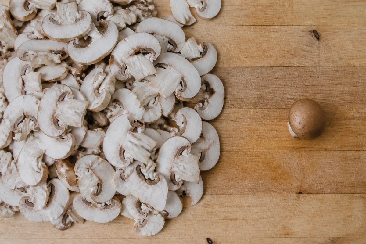 A wooden kitchen board with a large pile of cleaned sliced pale mushrooms and one small whole brown capped mushroom set apart on the bare board beside it