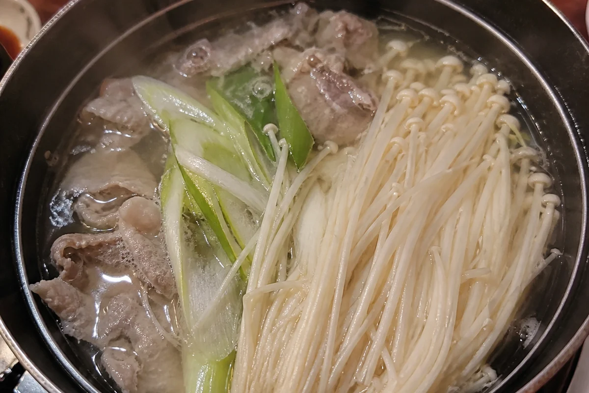 A simmering hot pot with clear broth, a bundle of long white enoki half submerged beside sliced spring onion and thin sliced meat, steam rising