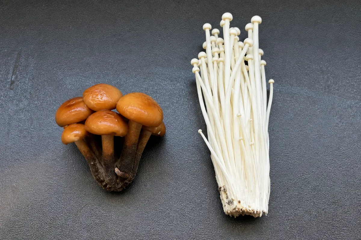 Two mushroom clusters side by side on a dark tray, the left a tight bunch of orange brown sticky capped mushrooms on short dark velvety stems and the right a tall bundle of long thin pure white stems with tiny caps