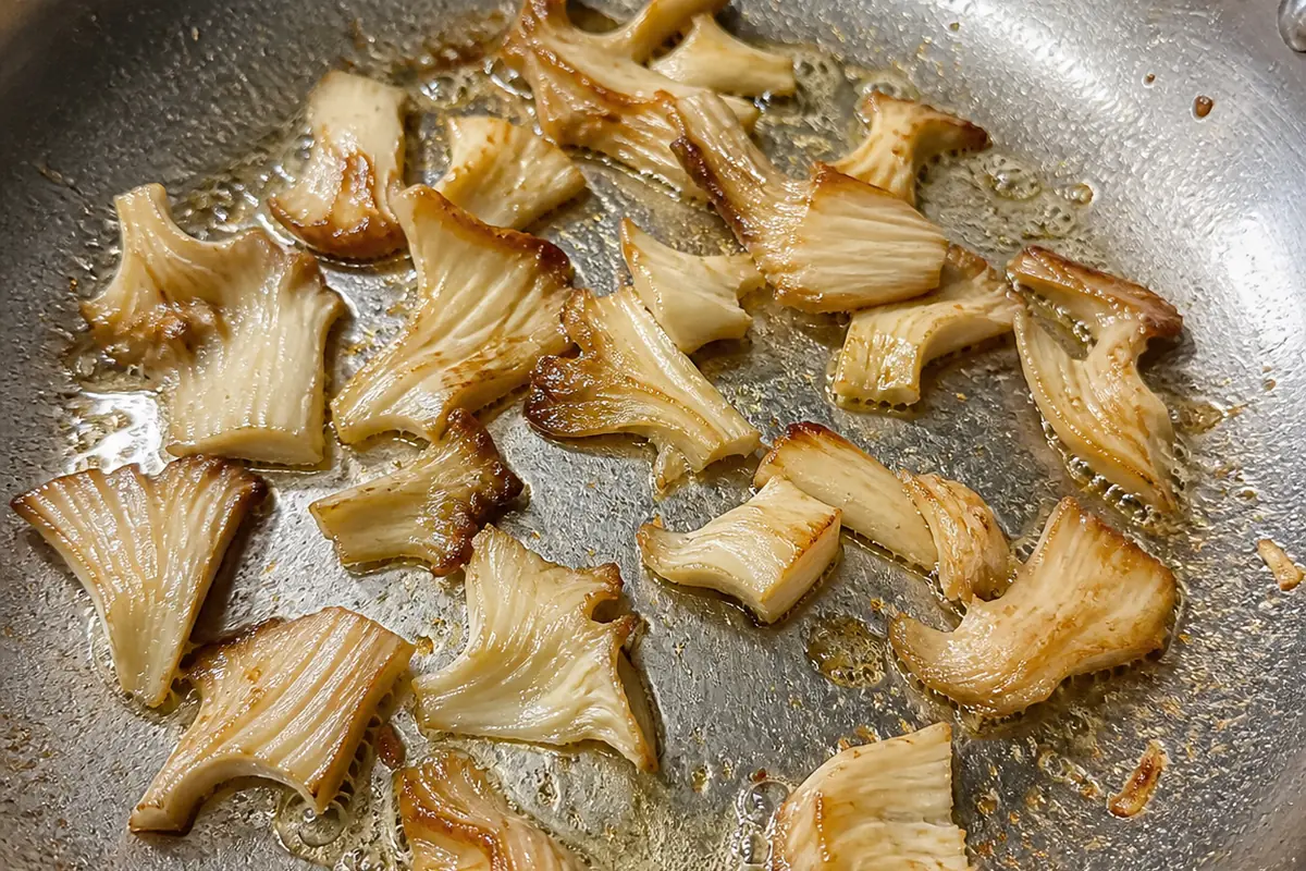 Torn pieces of pale mushroom frying in a stainless steel pan with butter, browned at the torn edges and holding their shape