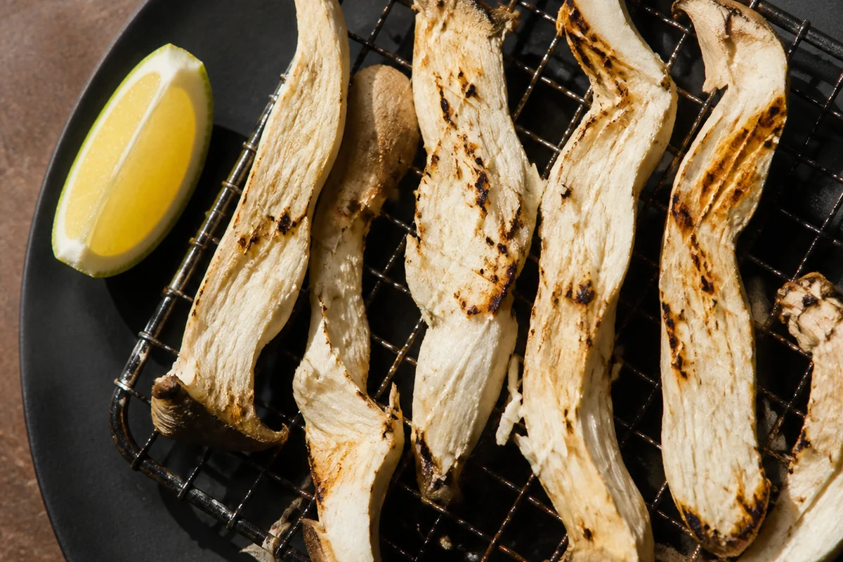 Thick hand-torn strips of pale mushroom grilling on a hot metal grate with light char marks and a wedge of citrus on a dark plate beside them