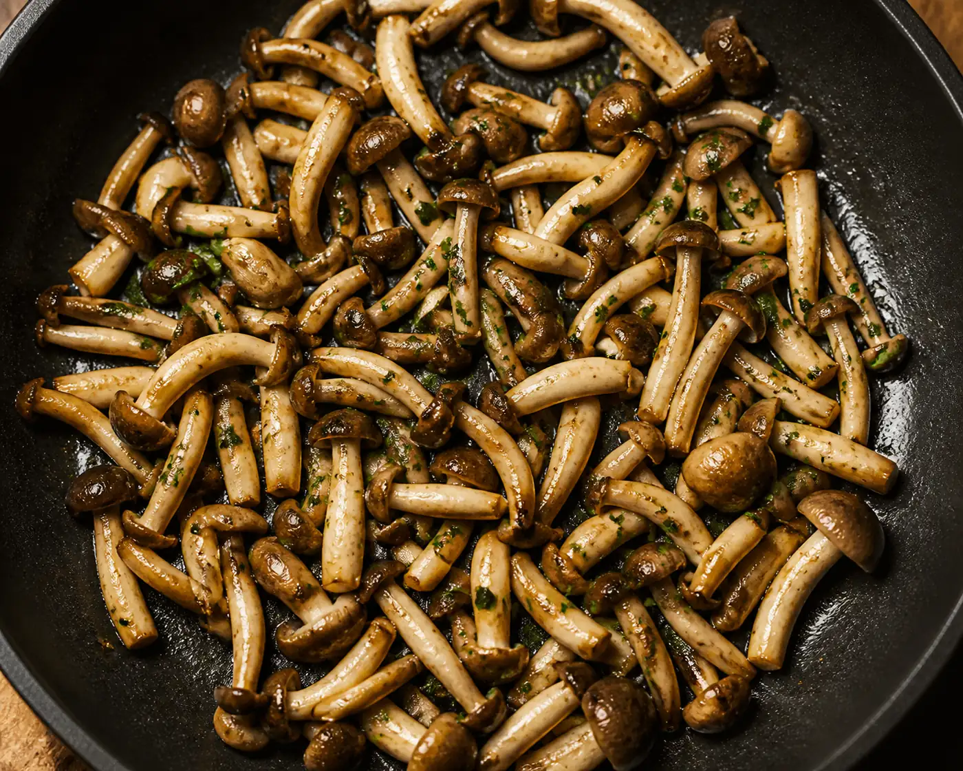 Finished sauteed beech mushrooms in a shallow black pan, browned and glossy with chopped green herbs scattered over them