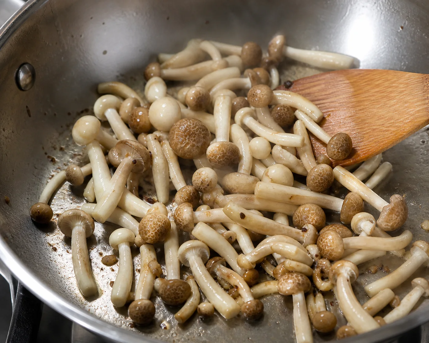 Separated beech mushrooms steaming in a hot steel pan with a wooden spatula resting in it, caps just starting to color at the edges