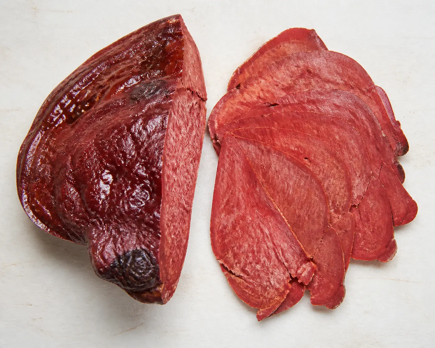 A thick red wedge of beefsteak fungus beside a fan of paper-thin translucent slices on a plain board