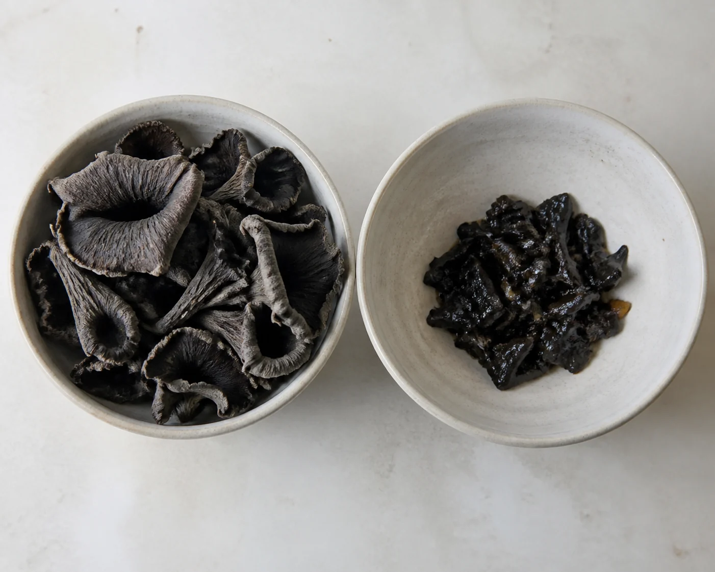 A raw handful of black trumpets in one shallow bowl beside the much smaller cooked portion in another