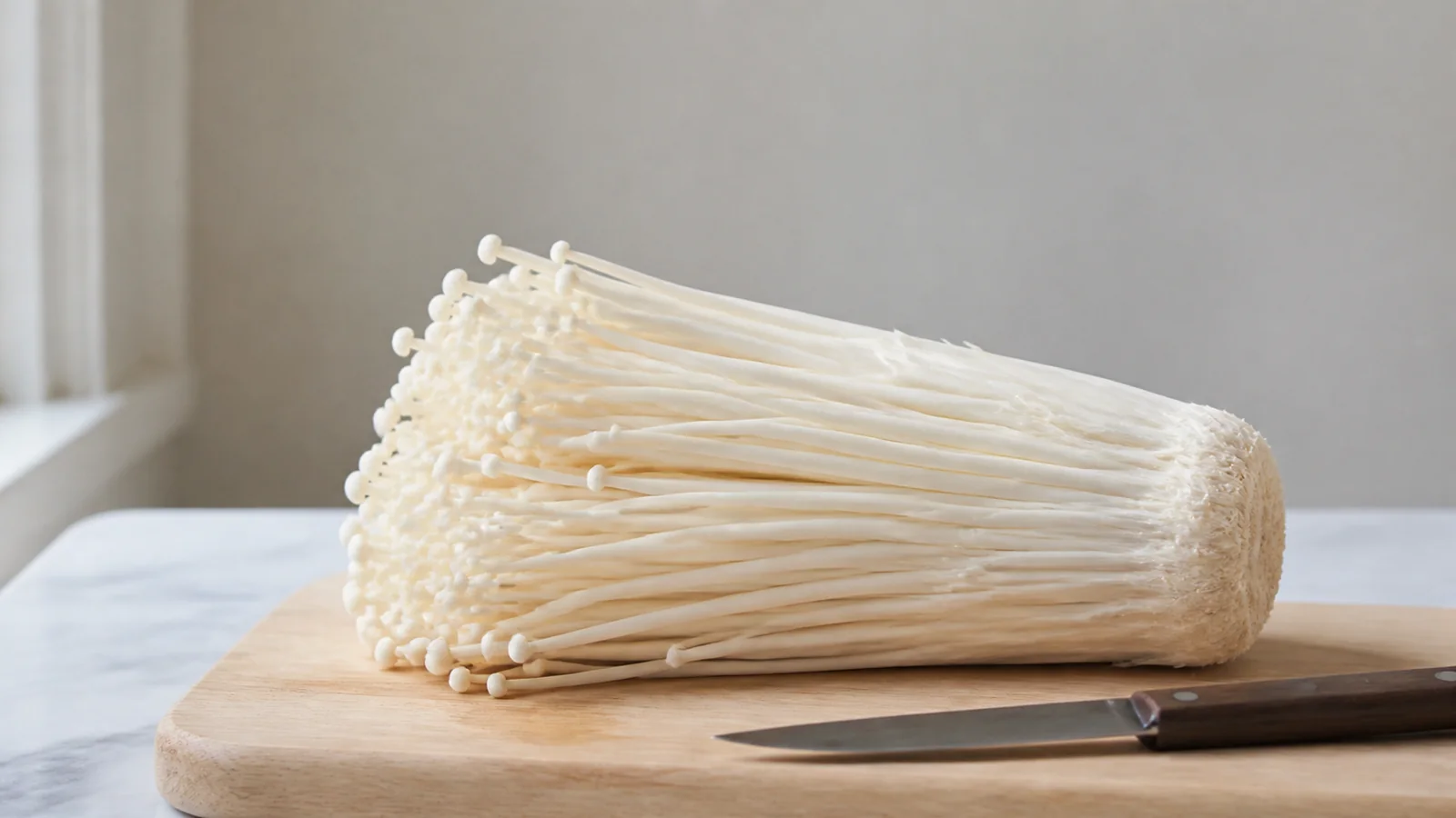 A fresh bundle of cultivated enoki with long white stems and tiny caps lying on a wooden kitchen board beside a knife