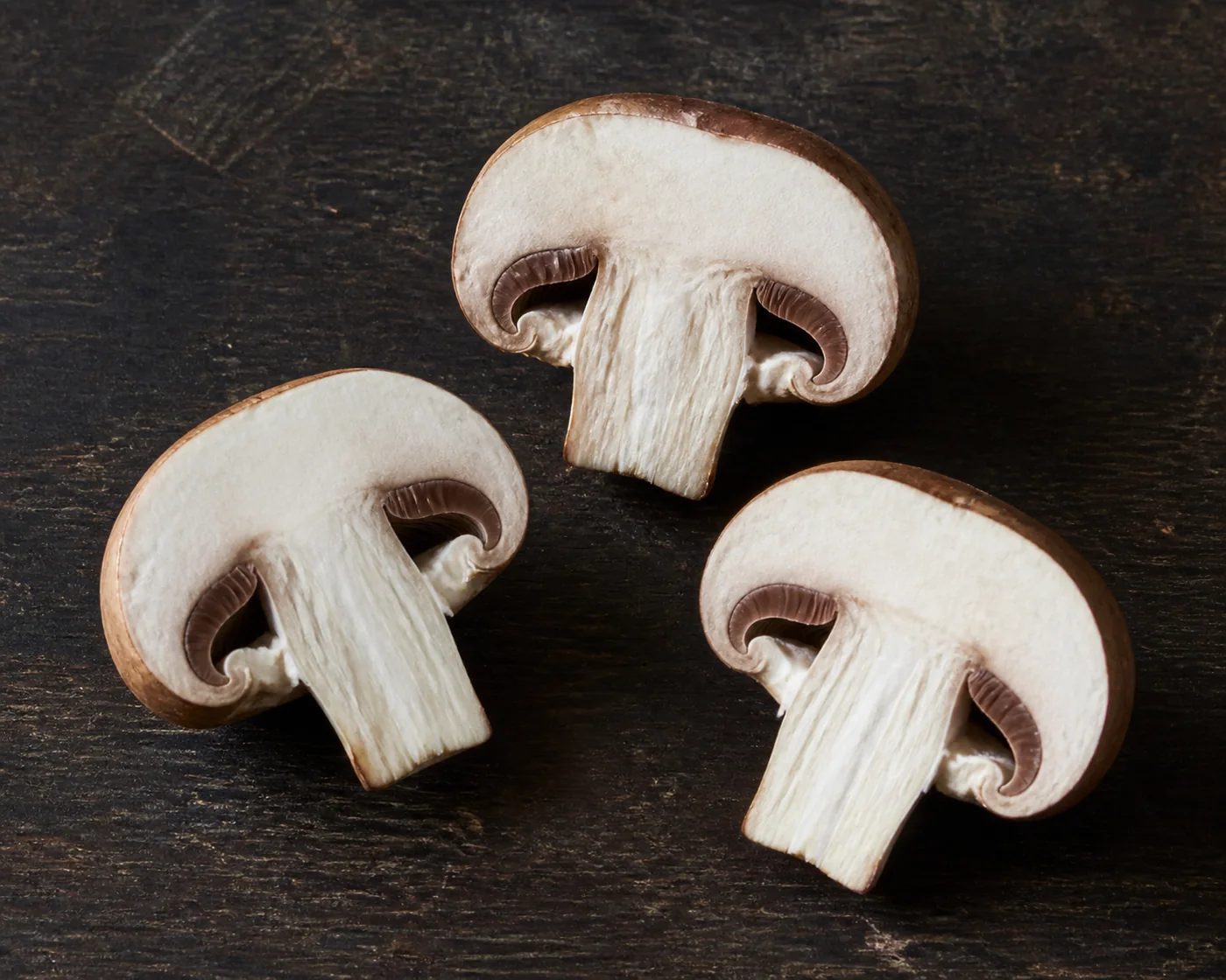Three cremini halved from cap to stem base on a dark board showing dense white flesh and a band of pink-brown gills