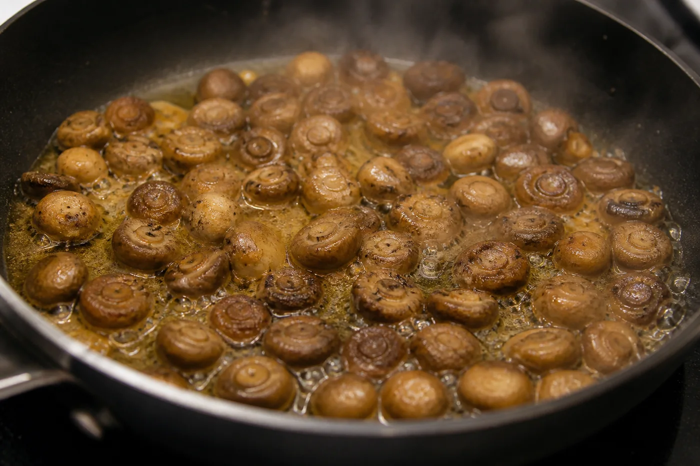 Many small tan mushroom caps simmering in a shallow pan of buttery golden liquid, well softened and darkened, with gentle steam rising