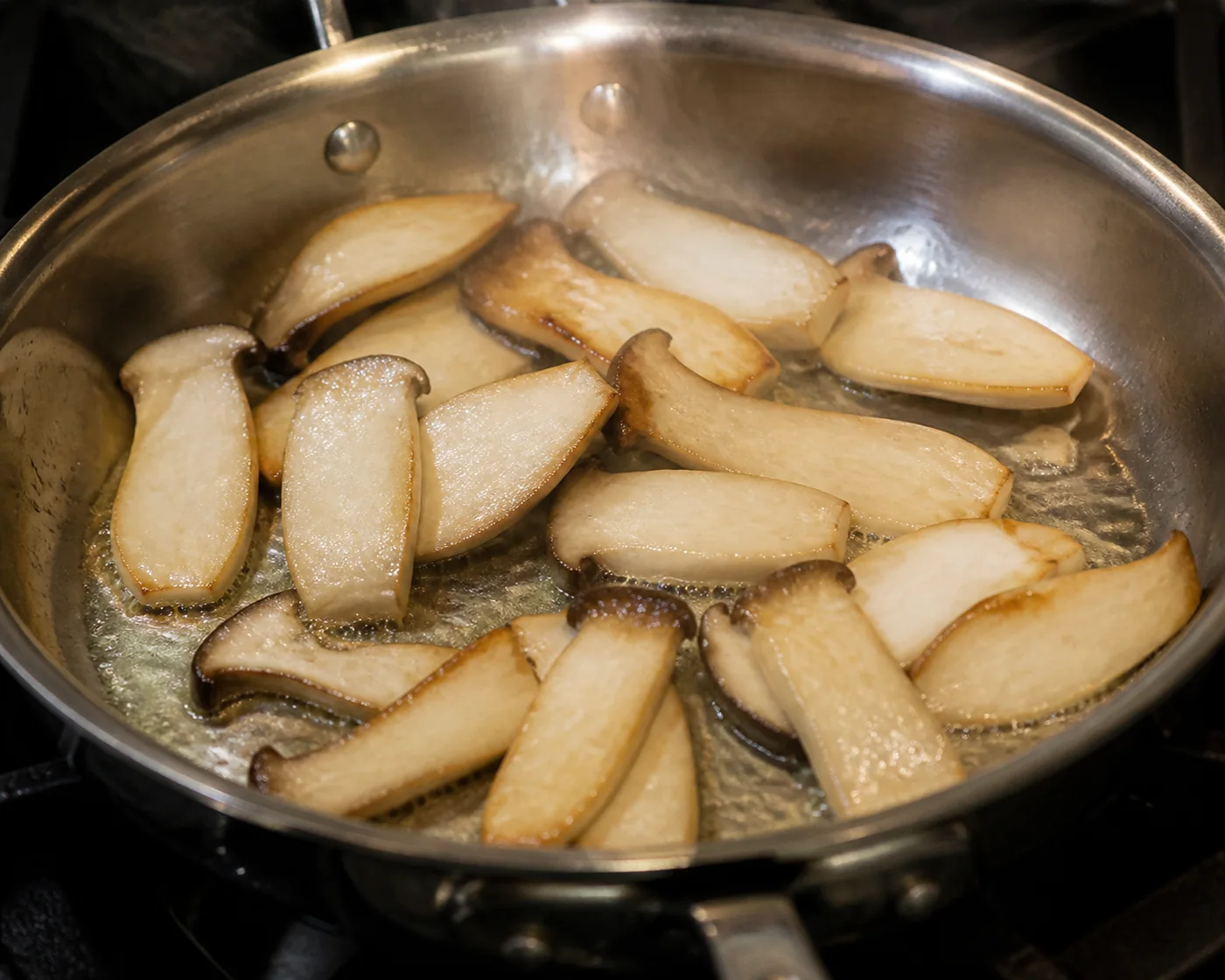 Thick pale mushroom slices sauteing in butter in a stainless steel pan with edges just beginning to brown and steam rising