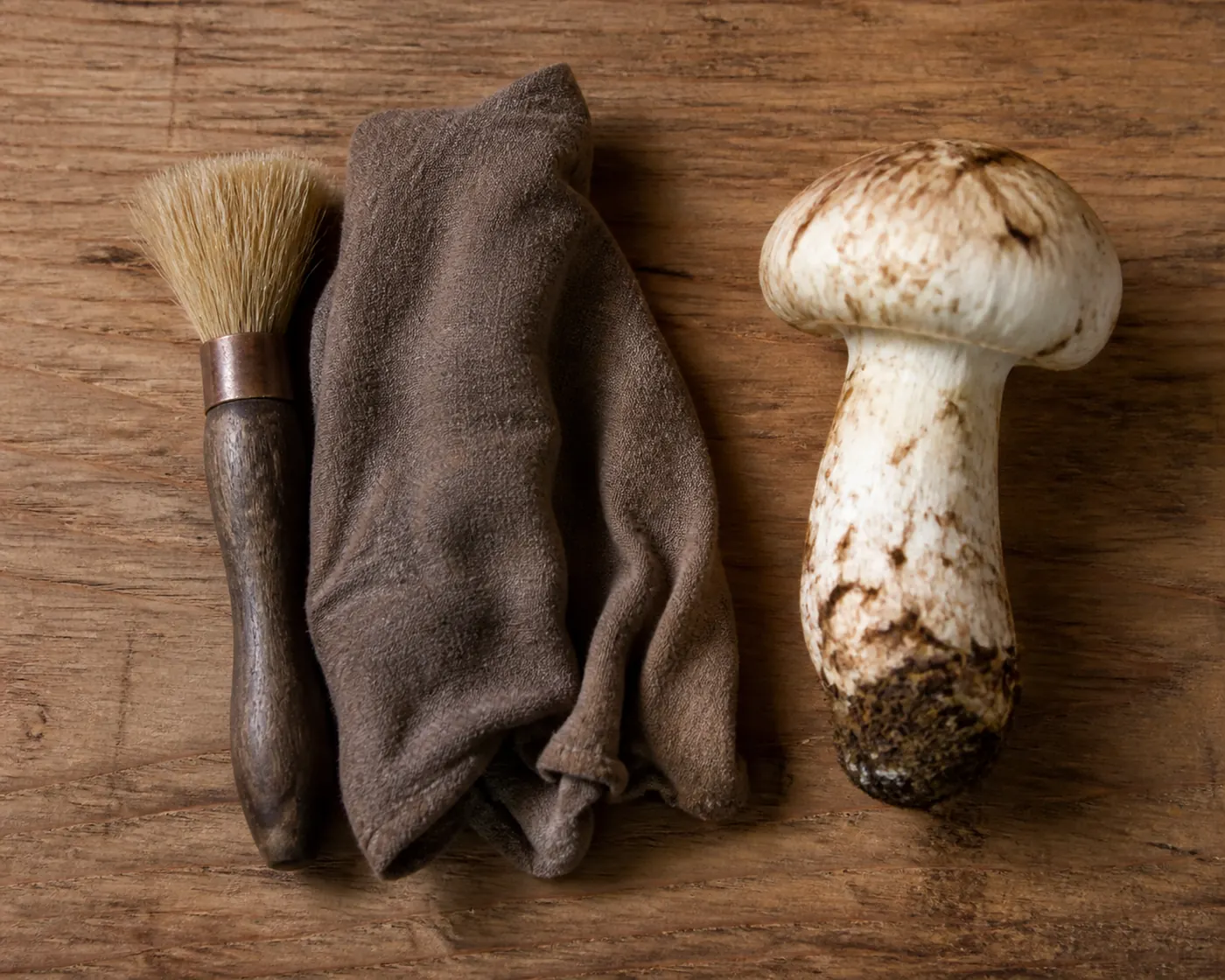 A soft-bristled mushroom brush and a folded damp cloth on a dry wooden board beside a whole matsutake with soil still on its base