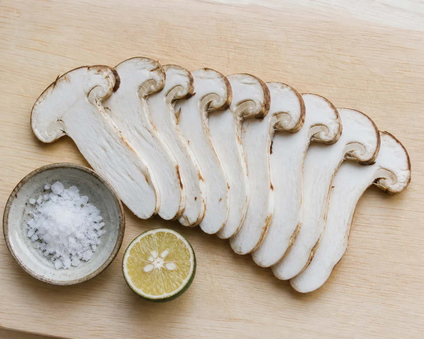 A matsutake mushroom sliced into very thin pale slices fanned along a wooden board with a small dish of flaky salt and a halved green citrus fruit beside them