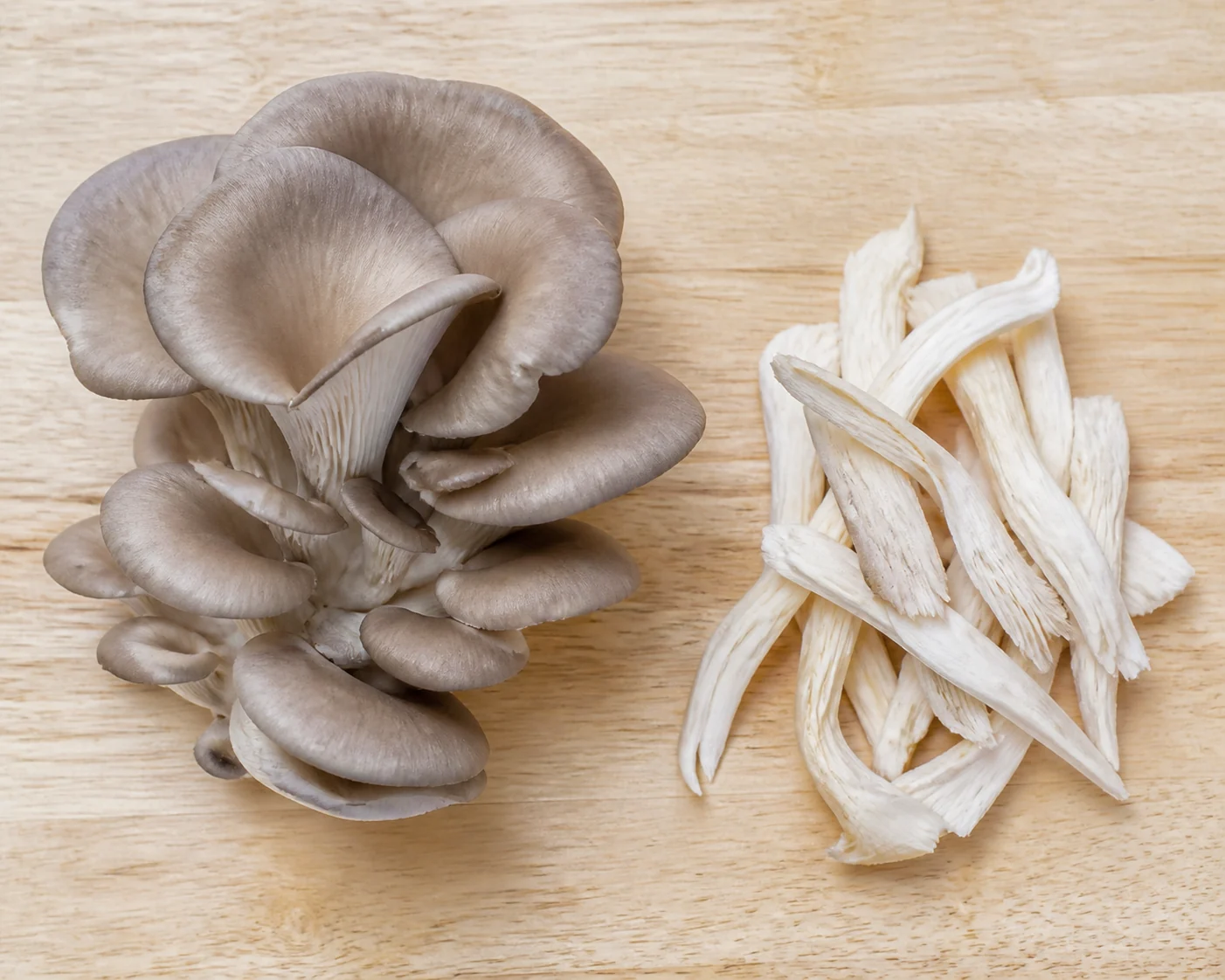 A cultivated oyster mushroom cluster on a pale board beside a small pile of thin hand-torn raw strips