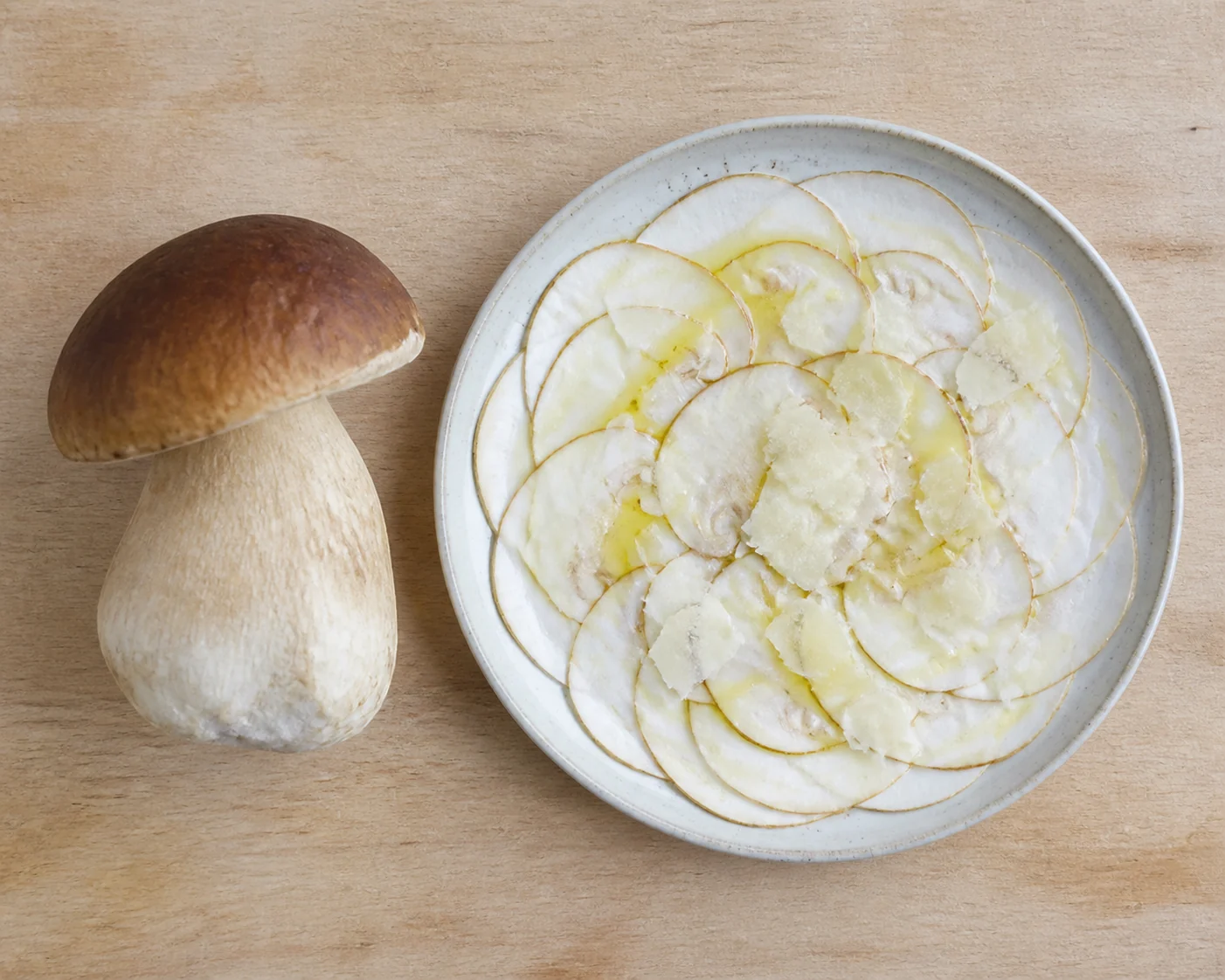One large firm porcini beside a plate of paper-thin raw slices dressed with oil on a pale board