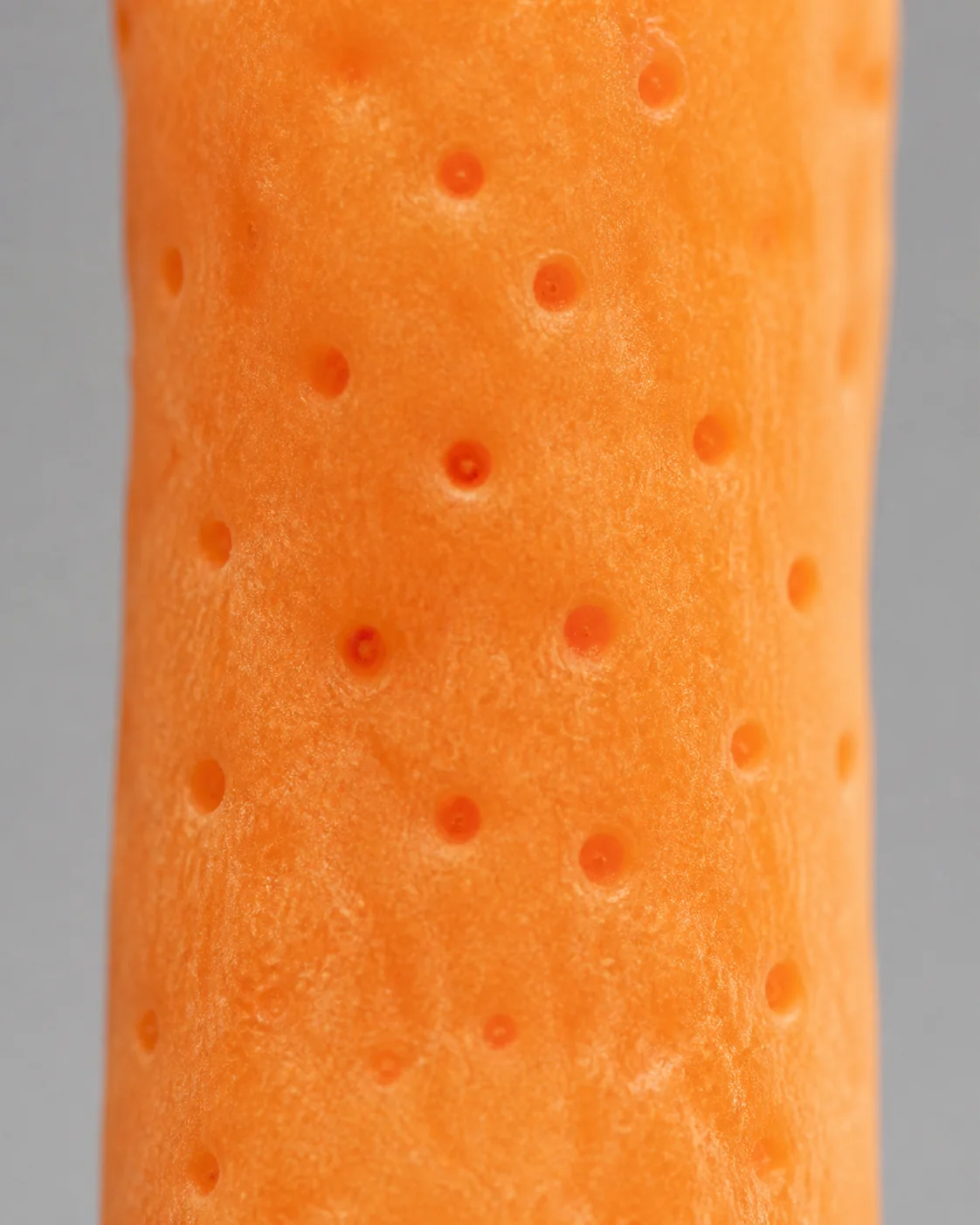 The stem of an orange mushroom showing scattered shallow circular dimples in a slightly darker orange, like small pits pressed into the surface